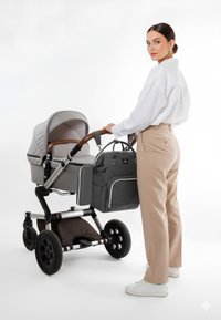 Grå stofbarnevogn med træhåndtag, store sorte hjul og en matchende grå pusletaske med quilteoverflade. Brugeren bærer beige bukser og en hvid skjorte.