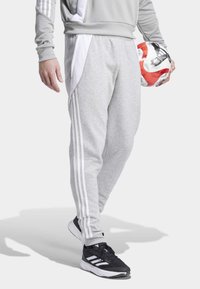 Pantalon de sport gris avec trois bandes blanches sur les côtés, associé à des chaussures de sport noires. Un ballon de football rouge et blanc est tenu à la main.