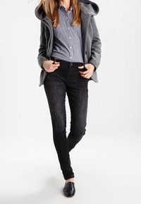 Grå huva kappa över en rutigt skjorta, kombinerad med mörka denimjeans och svart skot. Fokusera på överlappande texturer och lagerdesign.