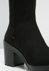 Bottine en daim noir avec un talon épais, une fermeture éclair latérale et une texture lisse, présentant un bout arrondi et des coutures renforcées.