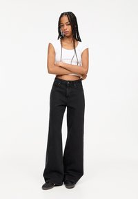 Schwarze weit geschnittene Jeans mit hoher Taille, aus Denimstoff. Kombiniert mit einem enganliegenden weißen Crop-Top. Das Design verfügt über die klassische Fünf-Taschen-Stil.