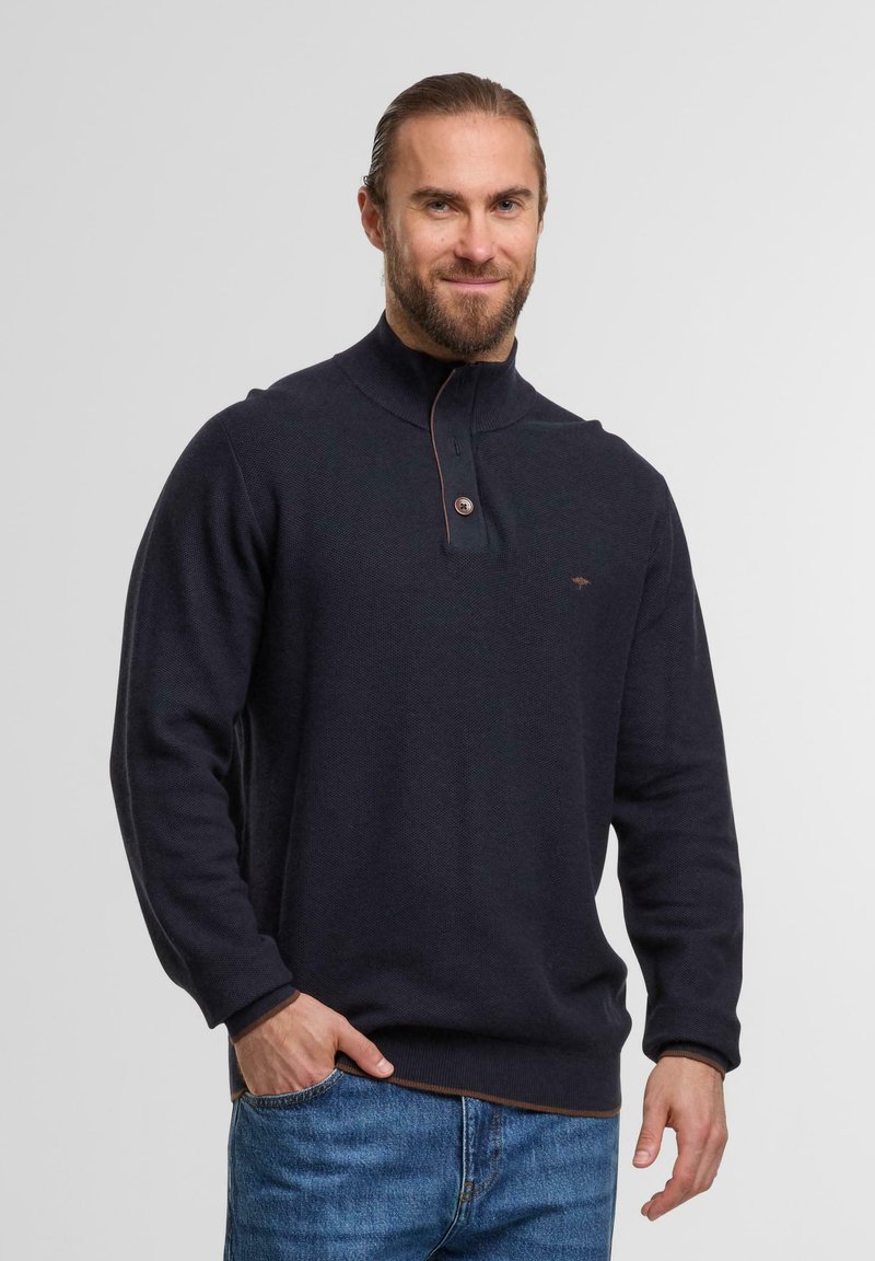 Mann mit Bart und zurückgebundenem Haar, der einen dunkelblauen, geknöpften Pullover und blaue Jeans trägt und mit einer Hand in der Tasche steht.