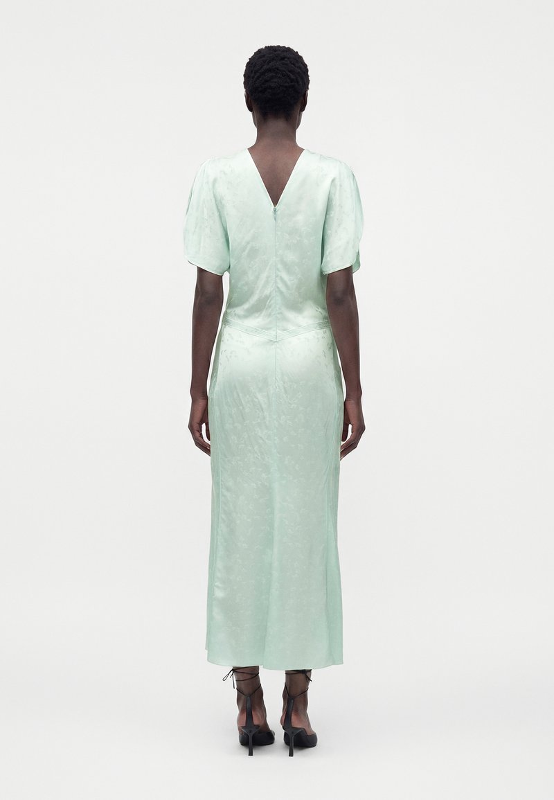 Femme aux cheveux courts et bouclés portant une robe midi vert pâle à manches bouffantes et des chaussures noires à talons hauts lacées, debout de dos.