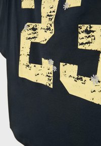 Sort stof med stort slidt gult nummer "23" trykt, dekoreret med små hvide broderede blomster spredt på og omkring tallene.