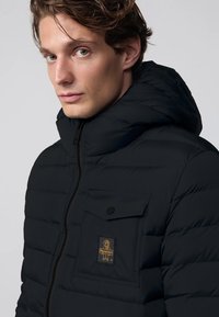 Giubbotto in piuma nero con cappuccio, tessuto testurizzato, chiusura frontale con zip e tasca sul petto con patch logo dorata.