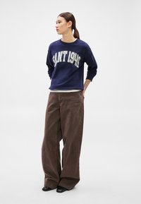 Tengerészkék pulóver "Saint 1942" felirattal, barna, széles szárú kordnadrággal párosítva. A fekete cipő teszi teljessé az összeállítást.