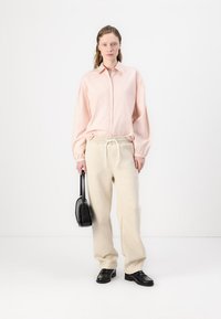 Light pink collared shirt with button closure, paired with cream sweatpants and black loafers. Model carries a small black handbag.