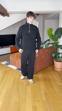 Felpa nera con zip e finitura testurizzata, abbinata a pantaloni neri dalla vestibilità ampia. Scarpe sportive argento, in piedi su pavimento in legno.