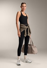 Černý sportovní crop top s bílým švem, černé legíny, béžový svetr uvázaný v pase, šedé tenisky a velká béžová sportovní taška.