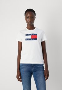 Biela bavlnená tričko s grafickým logom v modrej, červenej a bielej farbe, ktoré znie "TOMMY JEANS." Okrúhly výstrih a krátke rukávy.