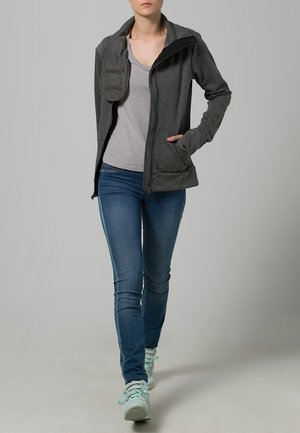 Veste grise à fermeture éclair avec col haut, poches latérales et tissu texturé. Portée sur un haut gris clair et associée à un jean bleu et des baskets claires.