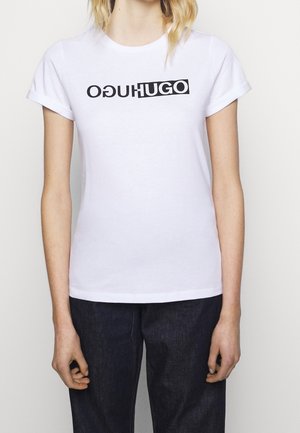 Osoba nosząca białą koszulkę z krótkim rękawem z czarnym tekstem "HUGO", częściowo odwróconym, połączoną z ciemnymi dżinsami, stojąca na jednolitym tle.