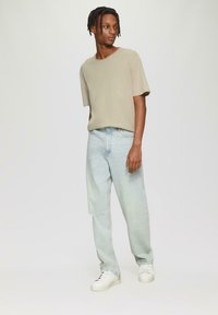 Lichtbeige T-shirt met korte mouwen en een ronde hals, gecombineerd met lichtblauwe tapered jeans. Model draagt witte sneakers. Eenvoudig, casual ontwerp.