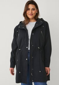 Zwarte capuchonjack met ritszakken, trekkoorden in de taille en knoopsluitingen. Gemaakt van een glad, lichtgewicht materiaal.