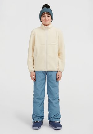 Fleece jacket in cream, zippered front, paired with blue snow pants and navy boots. Beanie with pom-pom completes the outfit.