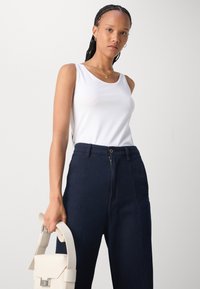 White sleeveless top paired with high-waisted dark blue trousers. Model holds a small, structured light beige handbag with a strap.