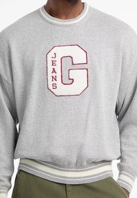 Maglione grigio con un logo ricamato "G" bianco e bordeaux e il testo "JEANS". Presenta un colletto e polsini a costine con accenti a righe.
