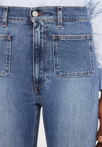 Visoko pasovi modri traper hlače z gladko teksturo, kontrastnim oranžnim šivom in dvema zadnjima žepoma s pravokotnimi pokrovi.