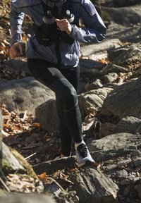 Svarta träningsleggar, grå lättviktjacka, springer på stenig terräng. Två vattenflaskor i ett bälte. Höstlöv syns.