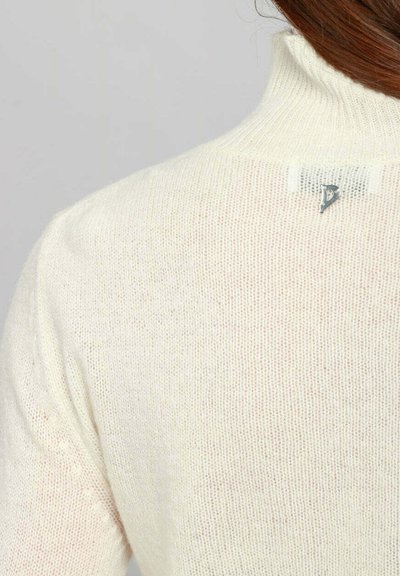 Primo piano di una persona che indossa un maglione a collo alto di colore crema, lavorato a maglia, con un piccolo logo metallico sul colletto posteriore.