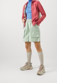 Persona con pantalones cortos de cargo verde claro, botas de senderismo beige con calcetas largas beige, camiseta azul y chaqueta roja sosteniendo la correa de una mochila.