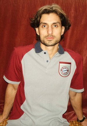 Jonge man met middellang bruin haar die een grijs en bordeauxrood FC Bayern München polohemd draagt tegen een rode achtergrond.