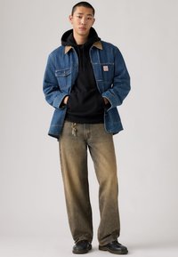 Τζιν μπουφάν με καφέ γιακά, μαύρο φούτερ από μέσα και ξεθωριασμένο μπεζ τζιν παντελόνι. Αξεσουάρ περιλαμβάνουν μπρελόκ στο τζιν και μαύρα παπούτσια.