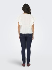 White blouse with ruffled sleeves, cinched waist, worn with dark skinny jeans and red flats featuring silver stud accents. Back view.