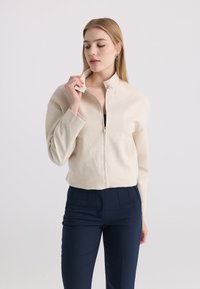 Femme aux longs cheveux blonds portant une veste crème zippée et un pantalon bleu marine, tenant le col de la veste avec la main gauche.