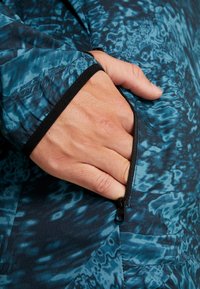 Détail de poche zippée sur un tissu à motifs bleu et bleu sarcelle foncé, présentant une texture lisse avec une bordure noire autour du poignet.