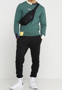 Felpa verde a maniche lunghe con logo, pantaloni sportivi neri, borsa a tracolla nera e scarpe da ginnastica bianche. Dettaglio sui polsini e sul colletto a coste.