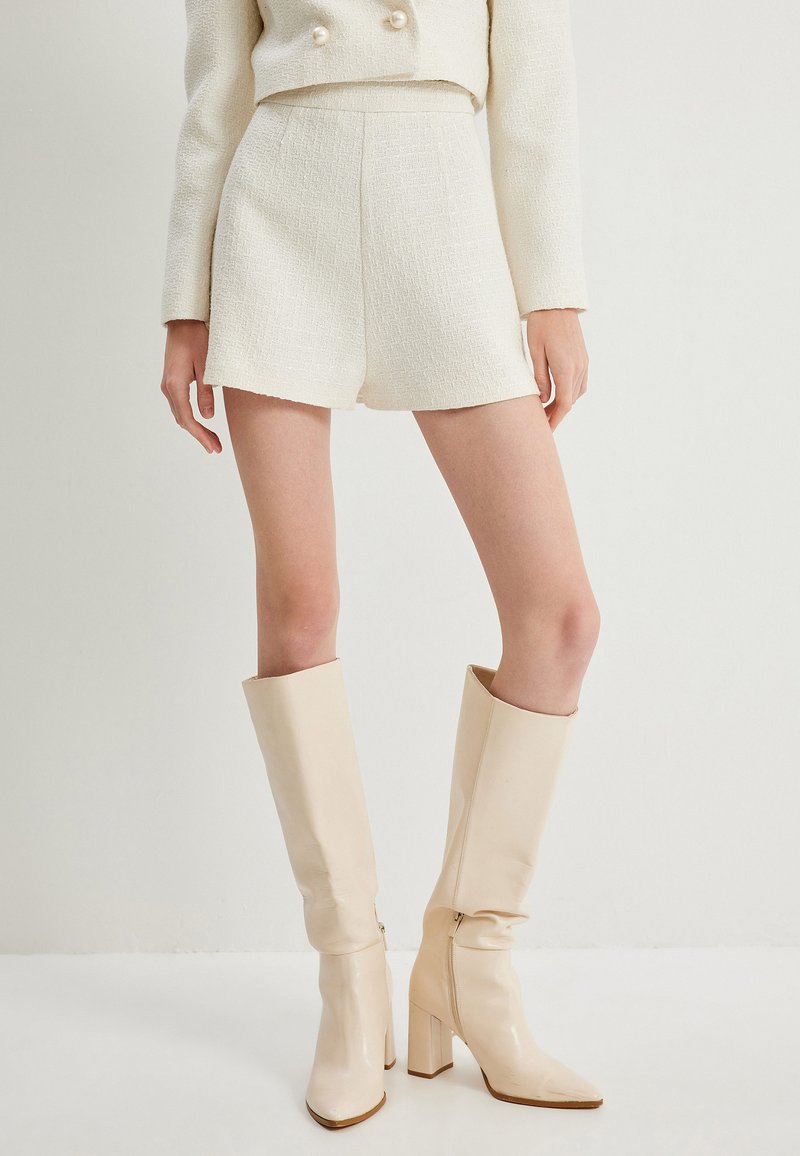 Witte, gestructureerde shorts gecombineerd met een cropped, dubbelgeknoopte jas met parelaccenten. Het model draagt glanzende beige kniehoge laarzen.