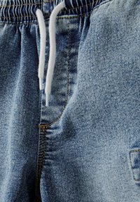 Denim joggerji s svetlo modrim pranjem, elastičnim pasom z nastavljivo vrvico, stranskim žepom in kontrastnim rumenim šivom.