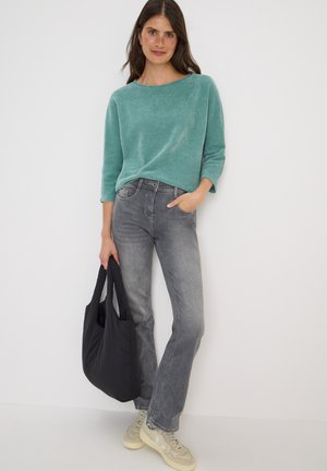 Pull en tricot turquoise à manches trois-quarts, associé à un jean gris clair et des baskets beiges. Le mannequin tient un sac fourre-tout noir.