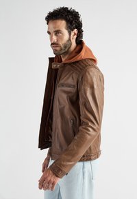 Bruine leren jas met een ritssluiting, voorzien van een geborduurd schouderontwerp, zijrits zakken en een contrasterende bruine hoodie eronder.