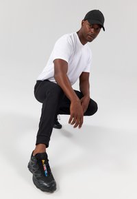 Witte t-shirt met korte mouwen, zwarte sportbroek en zwarte sneakers met gekleurde accenten. Het model staat in een hurkende positie tegen een neutrale achtergrond.