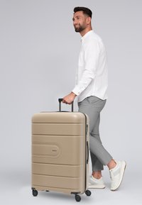 Harde koffer in beige met horizontale groeven, uitschuifbare handgreep en vier wielen, weergegeven naast een persoon in informele kleding.