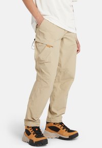 Beige cargobyxor med flera sidofickor och justerbara muddar, kombinerade med orangea och svarta vandringsskor med texturerade sulor.