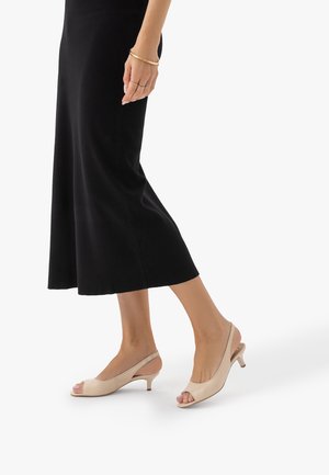 Kobieta w beżowych sandałkach na obcasie z odkrytymi palcami i czarnej spódnicy midi, stojąca z jedną stopą lekko wysuniętą do przodu na białym tle.