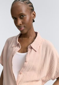 Camicia button-up di un rosa pallido, con texture e maniche corte, indossata sopra un top bianco, caratterizzata da una vestibilità rilassata e un tessuto morbido.