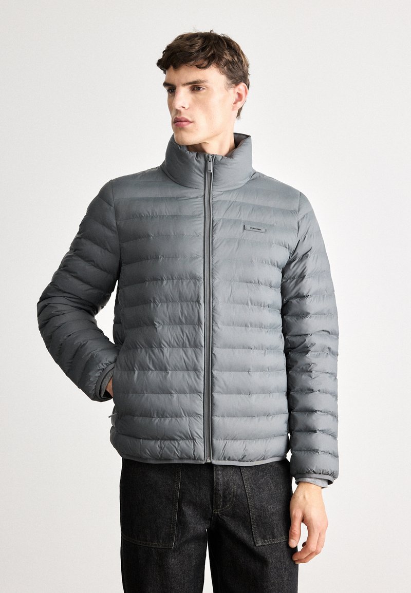 Pelēks pūkains mētelis ar augstu apkakli, horizontālu kvadrātu dizainu, priekšējo rāvējslēdzēju un logotipa uzkabi kreisajā pusē. Tekstūrēta auduma un pieguļoša silueta.