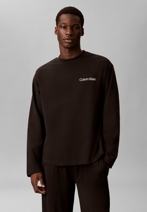 Junger Mann trägt ein schwarzes langärmeliges Calvin Klein Shirt und passende schwarze Hose vor einem einfarbigen grauen Hintergrund.