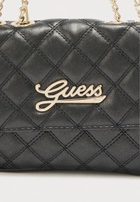 Μαύρη καπιτονέ τσάντα χειρός με γυαλιστερό φινίρισμα, χρυσό λογότυπο "Guess" και λουρί αλυσίδας με στρογγυλά εξαρτήματα.