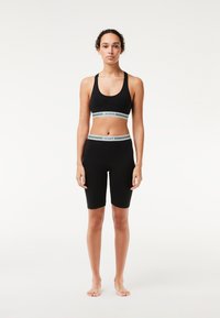 Set sportivo nero composto da un reggiseno sportivo aderente e pantaloni corti fino alla coscia, entrambi con bande del logo Lacoste in bianco e verde. Tessuto morbido e elasticizzato.