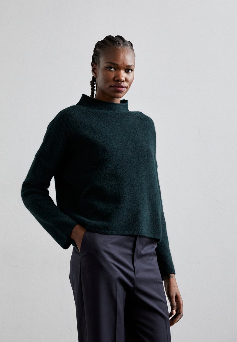 Maglione lavorato a maglia di un verde scuro, con un colletto alto e maniche lunghe, abbinato a pantaloni scuri, lisci e sartoriali. Fit sottile e rilassato.