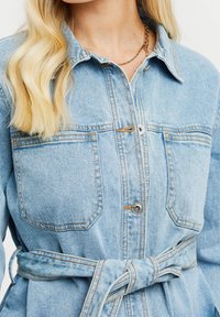 Vestido camisero de denim azul claro con cuello, botón en el frontal, dos bolsillos en el pecho y un cinturón atado en la cintura. Presenta costuras en contraste y una textura suave.