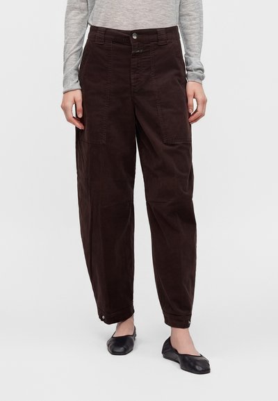 Personne portant un pantalon ample marron foncé avec de grandes poches à l'avant, un haut gris à manches longues et des chaussures plates noires, debout devant un fond uni.
