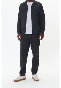 Czarna koszula na guziki nałożona na białą koszulkę, w połączeniu z czarnymi spodniami i białymi trampkami z brązowymi podeszwami. Gładki materiał, luźny krój.