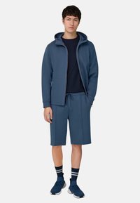Completo sportivo blu navy composto da una giacca con cappuccio e zip e pantaloncini abbinati, realizzato in un materiale liscio ed elastico.