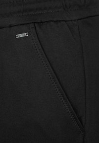 Pantalones negros con un bolsillo en la costura lateral con un detalle de puntada en zigzag y una pequeña etiqueta de metal cerca del borde del bolsillo. Textura de tela suave.